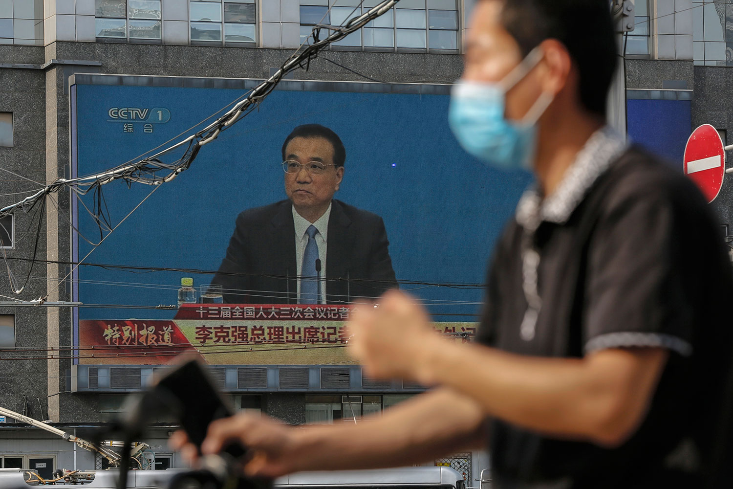 图为，2020年5月28日，一名戴着自行车防护面罩的男子在大屏幕附近经过，屏幕显示中国国务院总理李克强在人大闭幕后的中外记者会上。（美联社）